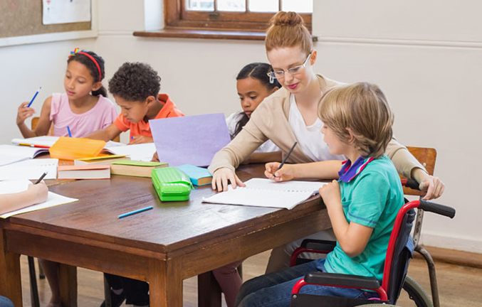 Young students sitting at wooden desk in classroom; teacher helping boy in wheelchair