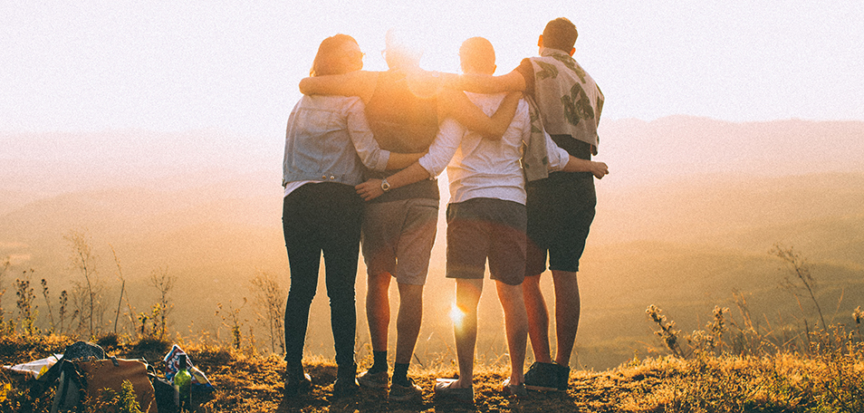 Four young adults stand shoulder-to-shoulder with their arms around each other overlooking a valley. Sun shines in distance.